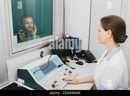 Audiologin Frau, die die Höruntersuchung bei einem Mann mit gemischter Rasse mit einem Audiometer in einem speziellen Audioraum macht. Stockfoto