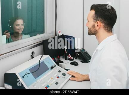 Audiologe Mann, der die Höruntersuchung bei einer erwachsenen Patientin mit einem Audiometer in einem speziellen Audioraum macht. Stockfoto