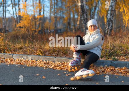 Ein Mädchen in Rollschuhe sitzt auf Betonkandare im Park und schaut weg. Eine junge Eiskunstläuferin ruht. Herbstabend. Das Mädchen trägt einen weißen Hut, Pullover, Jacke und schwarze Leggings. Speicherplatz kopieren. Stockfoto