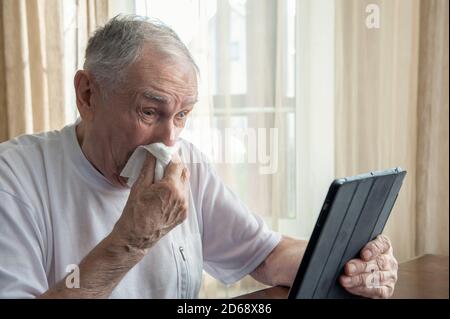 Ältere Menschen mit Allergien und Erkältungen hustet und niest in eine Serviette. Ein kranker alter Mann kauft online während der Pandemie zu Hause Stockfoto