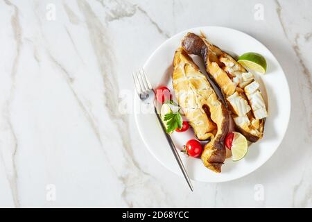 Traditionelle Alaskan kalt geräucherte Heilbutt Fisch Steaks mit Limette serviert Mit Rosmarinzweigen und Tomaten auf einem Teller mit Gabel Auf einem weißen Marmorhintergrund Stockfoto