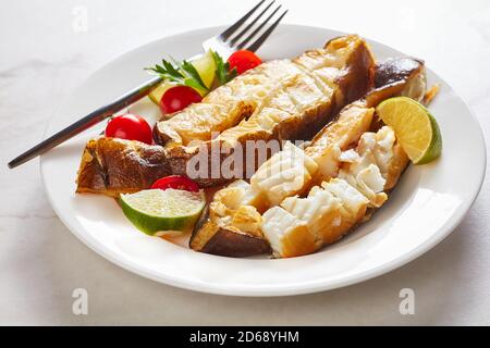 Traditionelle Alaskan kalt geräucherte Heilbutt Fisch Steaks mit Limette serviert Mit Rosmarinzweigen und Tomaten auf einem Teller mit Gabel Auf einem weißen Marmorhintergrund Stockfoto