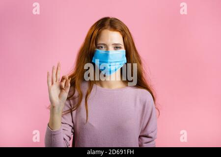 Glückliche Frau in medizinische Maske auf rosa Hintergrund. Lady mit langen roten Haaren zeigt OK Geste. Gewinner. Erfolg. Körpersprache. Stockfoto