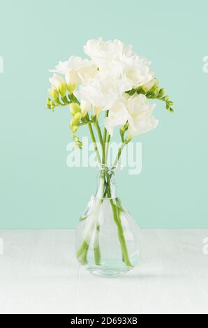Romantische Frühlingsblumen Freesia in durchsichtiger Glasvase in grüner Minze menthe Innenraum auf weißem Holzbrett, vertikal. Stockfoto