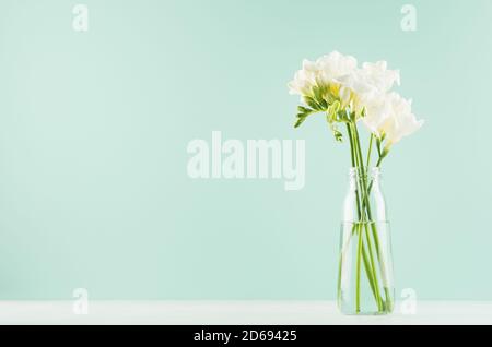 Duft weiche helle weiße Blumen Freesia in Bouquet in einfache Glasflasche in grüner Minze menthe Interieur auf weißem Holzbrett. Stockfoto