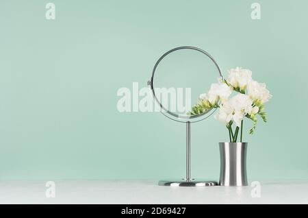 Elegante Frisierkommode mit weißen Blüten Freesie und runden Spiegel auf weißem Tisch und grünen Minze menthe Wand. Stockfoto