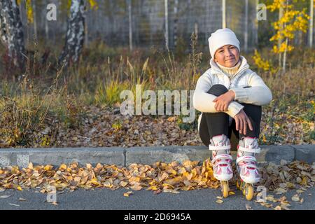 Ein Mädchen in Rollschuhe sitzt auf Beton Bordstein im Park. Eine junge Eiskunstläuferin ruht. Herbstabend. Das Mädchen trägt einen weißen Hut, Pullover, Jacke und schwarze Leggings. Speicherplatz kopieren. Stockfoto