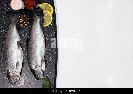 Frische Regenbogenforellen auf dem schwarzen Brett auf dem Tisch mit Salz, Pfeffer, Zitrone und Chili-Pfeffer Zutaten Stockfoto