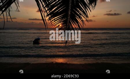 Schöner friedlicher Sonnenuntergang am tropischen Strand Anse Forbans im Süden der Insel Mahe, Seychellen mit Sonne scheint durch Palmblatt, Boot Docking. Stockfoto