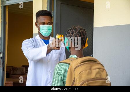 Lehrerin in einer afrikanischen Schule, die die Temperatur der Schülerin vor ihr überprüft Tritt in den Schulungsraum ein Stockfoto