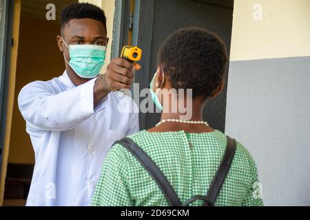 Männlicher Lehrer in einer afrikanischen Schule, der den Körper eines Schülers überprüft Temperatur mit einem Infrarot-Thermometer, bevor sie in den Klassenzimmer Stockfoto