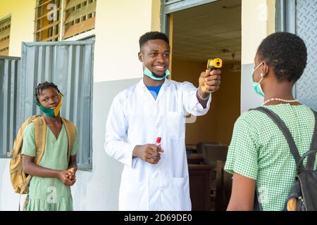 Lehrer in einer afrikanischen Grundschule Überprüfung der Temperatur des Schulkindes Bevor sie das Klassenzimmer betritt Stockfoto