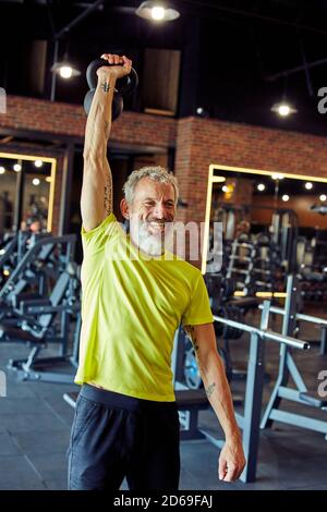 Vertikale Aufnahme eines lächelnden reifen kaukasischen Mannes in Sportkleidung Training mit Kurzhanteln im Fitnessstudio Stockfoto