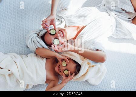 Junge positive Mutter und Tochter mit Handtuch auf dem Kopf liegend Auf dem Bett zusammen mit Gurke Stockfoto