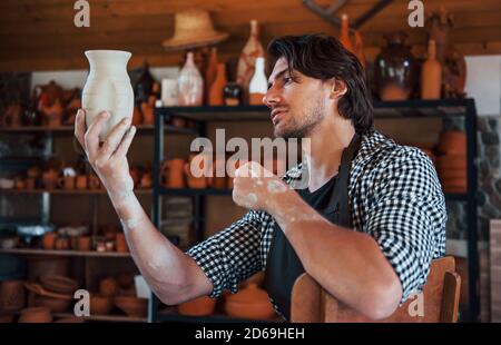 Junger Keramiker hält frischen handgemachten Topf in der Hand und sieht aus Bei den Ergebnissen seiner Arbeit Stockfoto