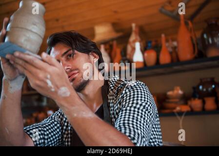 Junger Keramiker hält frischen handgemachten Topf in der Hand und sieht aus Bei den Ergebnissen seiner Arbeit Stockfoto