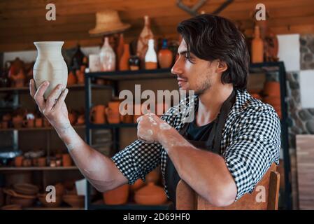 Junger Keramiker hält frischen handgemachten Topf in der Hand und sieht aus Bei den Ergebnissen seiner Arbeit Stockfoto