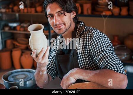 Junger Keramiker hält frischen handgemachten Topf in der Hand. Zufrieden mit den Ergebnissen seiner Arbeit Stockfoto