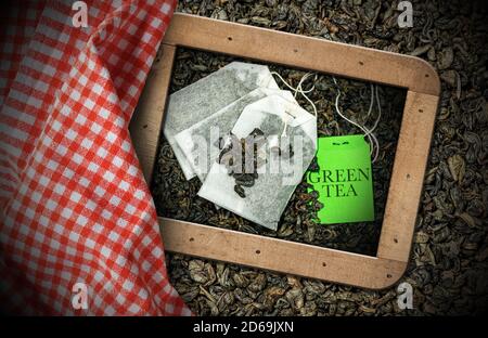 Grüner Teebeutel in einem Holzrahmen mit getrockneten Blättern auf dem Hintergrund, und teilweise von einer rot-weiß karierten Tischdecke bedeckt Stockfoto
