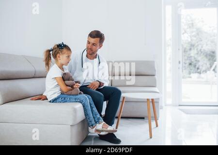 Männlicher Arzt in weißer Uniform sitzt in der Klinik mit Kleines Mädchen Stockfoto