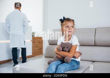 Ärzte dahinter. Nettes kleines Mädchen mit Teddybär in den Händen sitzt im Wartezimmer des Krankenhauses Stockfoto