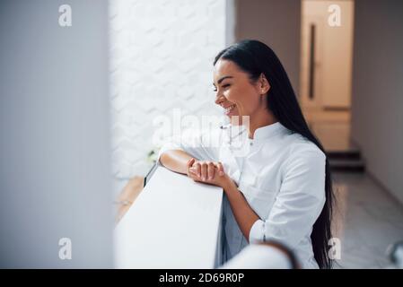 Positiv lächelnde Krankenschwester steht in der Rezeption der Klinik und Sich auf den Tisch lehnen Stockfoto