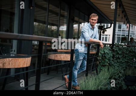 Auf das Geländer gelehnt. Vorderansicht eines Typen in Jeans, der tagsüber im Café steht Stockfoto