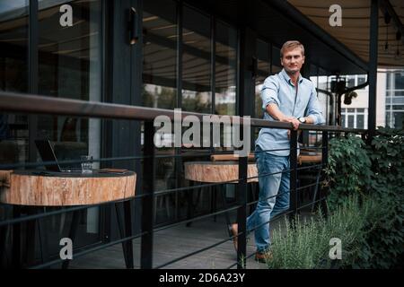 Auf das Geländer gelehnt. Vorderansicht eines Typen in Jeans, der tagsüber im Café steht Stockfoto