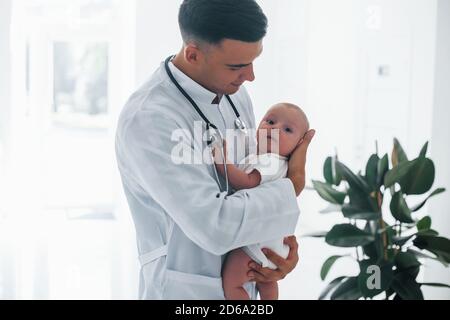 Steht gegen das Fenster. Der junge Kinderarzt ist mit dem kleinen Baby tagsüber in der Klinik Stockfoto