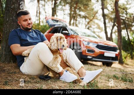 Der Mann sitzt in der Nähe des Baumes mit seinem Hund im Freien Der Wald Stockfoto