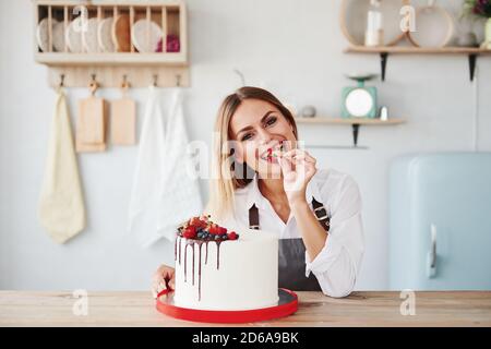 Positive Blondine in drinnen mit ihrem hausgemachten Kuchen hält Früchte Stockfoto