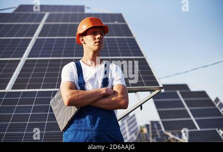 Steht mit gekreuzten Armen. Männlicher Arbeiter in blauer Uniform im Freien mit Solarbatterien an sonnigen Tagen Stockfoto