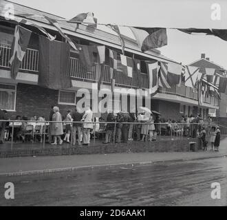 1970er Jahre, historisch, an einem regnerischen nassen Tag, draußen auf dem überdachten Gang eines Blocks von gemeinderatswohnungen, in Lewisham, Southeast London, England, Großbritannien, Kinder, die an langen Tischen sitzen, um eine Party mit Eltern zu feiern. Stockfoto