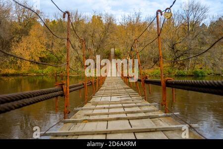 Kleine Hängebrücke über den Fluss im Herbstwald. Hängebrücke im Freien aus Holz und Metall. Geschlossene Vorhängeschlösser, die als Symbol ewiger Liebe an der Geländer hängen. Stockfoto