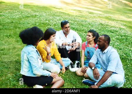 Afghanischer, schwarzer, mexikanischer und indischer Plaudernd, während er im Park auf dem Gras sitzt. Das Konzept der Kommunikation verschiedener Nationalitäten. Stockfoto