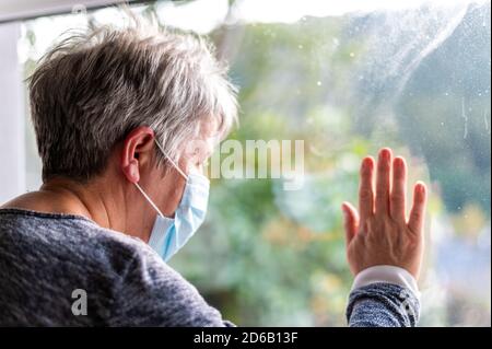 Ältere Frau trägt eine schützende Gesichtsmaske und blickt aus dem Fenster, mit Covid-Virus-Zellen außerhalb des Fensters. Stay Safe Stay Home Konzept. Flacher Fokus auf Stockfoto