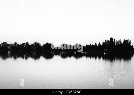 Fantastische Graustufenaufnahme einer kleinen Insel mit Bäumen, die sich spiegeln Auf einem ruhigen ruhigen See Wasser Stockfoto