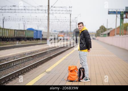 Der junge Mann steht auf dem Bahnsteig und wartet auf den Zug. Männlicher Passagier mit Rucksäcken auf Bahnsteig in Warten auf Zugfahrt. Konzept des Tourismus Stockfoto