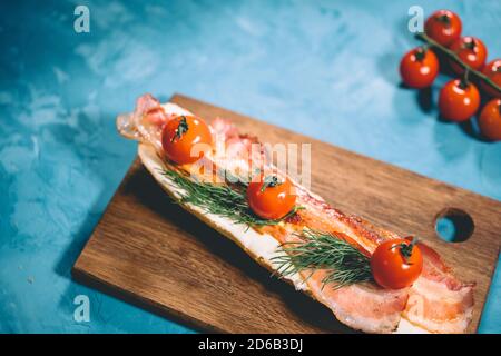 Stillleben von appetitanregenden Bruschetta auf Holzbrett. Nahaufnahme von Sandwich mit geschmolzenem Käse, Speck, Kirschtomaten und Dill auf blauem Hintergrund. Stockfoto