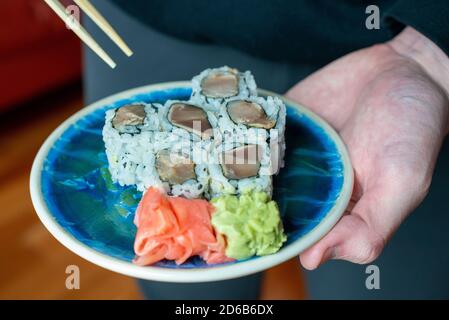 Ein kleiner blauer Porzellanteller mit Sushi und hölzernen Essstäbchen, die in der Hand eines Mannes gehalten werden. Stockfoto