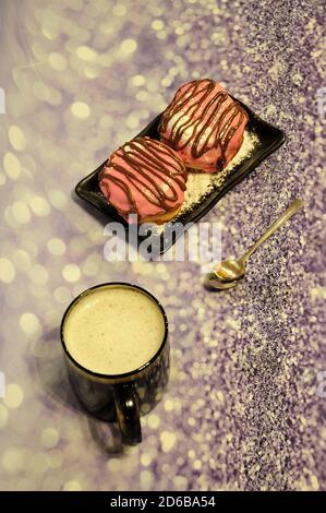 Ein Becher mit frischem Cappuccino mit Schaum und ein Teller mit zwei Donuts in rosa Glasur mit Schokoladendekor auf grauem Glitzer-Hintergrund. Nahaufnahme. Stockfoto