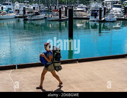 Airlie Beach, Queensland, Australien - März 2020: Eine Backpacker-Frau, die neben dem Hafen von Airlie Marina spazierengeht und mit ihrer Reiseausrüstung beladen ist Stockfoto