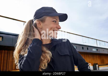 Porträt einer lächelnden Frau in Arbeitsuniform und Mütze in der Nähe des Gebäudes. Sieht aus, als würde sie im Lieferlager oder im professionellen technischen Kundendienst arbeiten Stockfoto