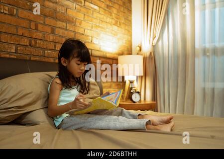 Kleines Mädchen in grünen Pyjamas halten Buch, suchen das Buch, während sie in ihrem Schlafzimmer sitzen Stockfoto