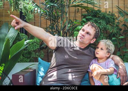 Vater blonde Sohn sitzen Blick weit weg, Vater zeigen mit Finger. Papa umarmt sanft Jungen Kind. Helle Sonnenstrahlen Licht verlässt Gartenterrasse. Frohe Kindheit Stockfoto