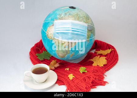 Der Globus trägt eine medizinische Maske und rot warmen Schal mit Ahornblättern und einer Tasse Tee. Weißer Hintergrund. Konzept, unsere Welt ist mit Coronavirus infiziert. Herbst, zu Hause in Quarantäne bleiben. Hochwertige Fotos Stockfoto