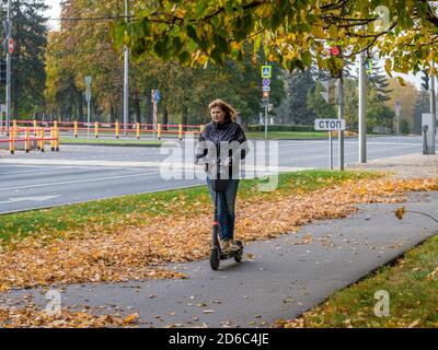 Moskau. Russland. 11. Oktober 2020. Junge Frau fährt einen Elektroroller auf dem Bürgersteig in einem Stadtpark. Spaziergang auf der Straße an einem Herbsttag. Es gibt Stockfoto