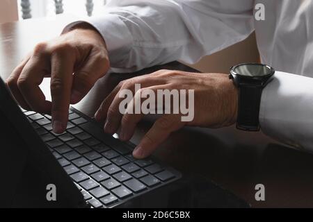 Nahaufnahme der Hände eines Mannes in einem Hemd, der auf der Tastatur eines Tablets oder Laptops tippt. Am Handgelenk trägt sie eine Smartwatch und ein Smartphone in der BA Stockfoto