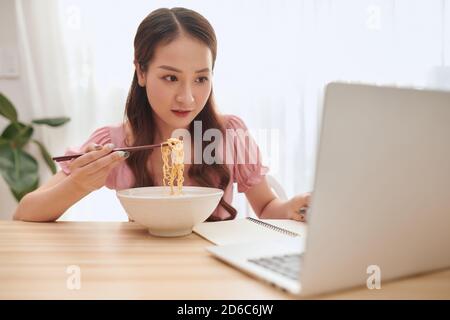 Schöne junge asiatische Frau mit Laptop und asiatische Nudeln zu Hause essen. Stockfoto