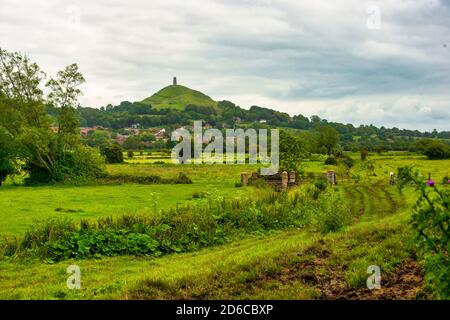 Glastonbury Tor ragt über das Township Glastonbury, mit rauem Ackerland im Vordergrund. Kleine Steinbrücke im Vordergrund. Stockfoto